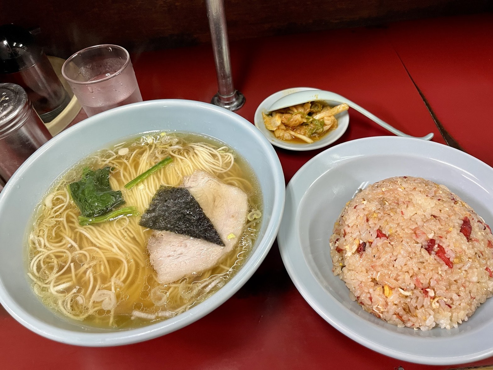 赤テーブルに置かれた大きめ丼の醤油ラーメンと、隣に盛られたピンクのチャーハン、奥に小皿のおかず